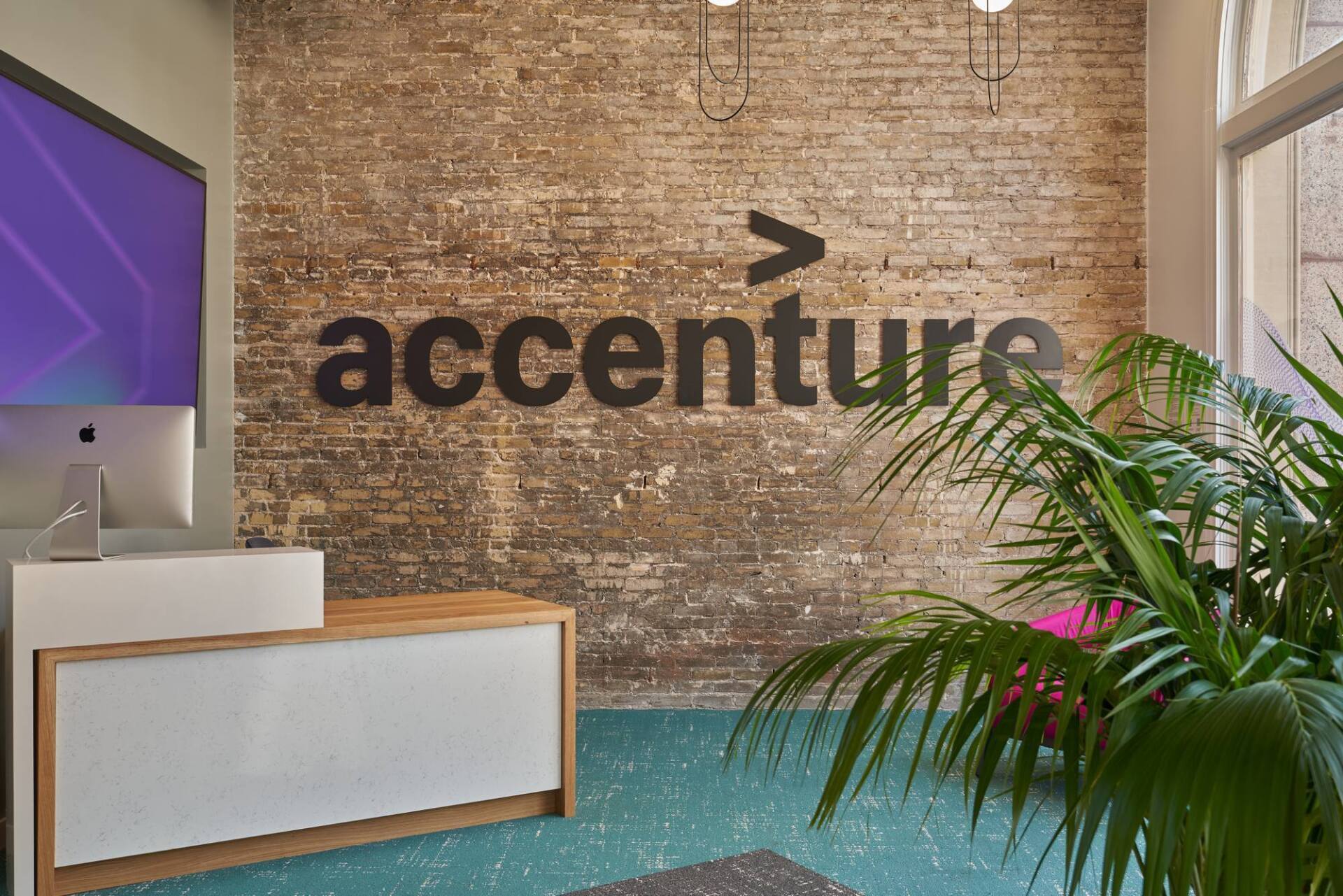 Accenture logo on a brick wall above a reception desk with a plant in the corner.