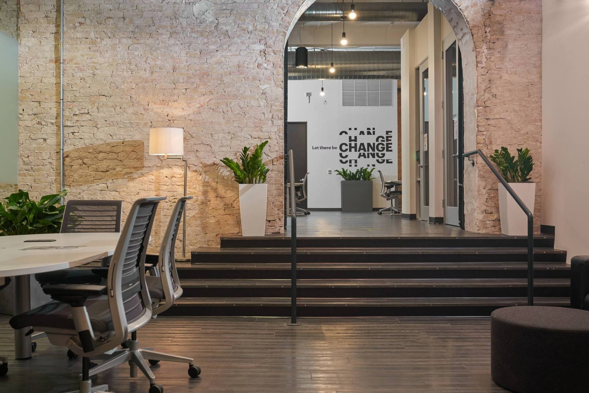 Office interior with stairs leading to a doorway, plants, a brick wall, chairs, and a table.