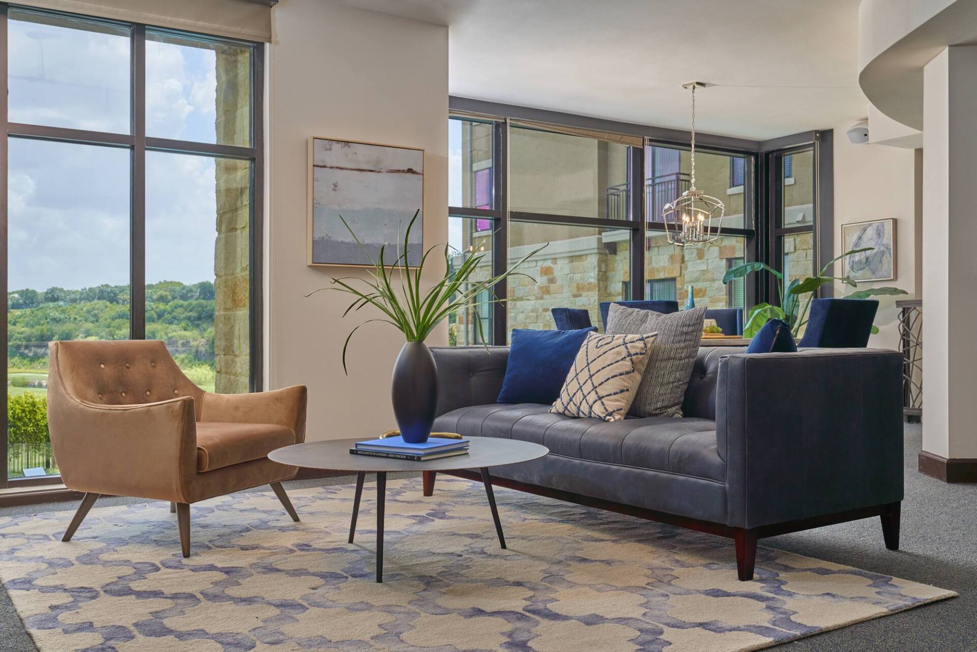 Modern living room with blue sofa, tan armchair, area rug, and large windows overlooking a green landscape.
