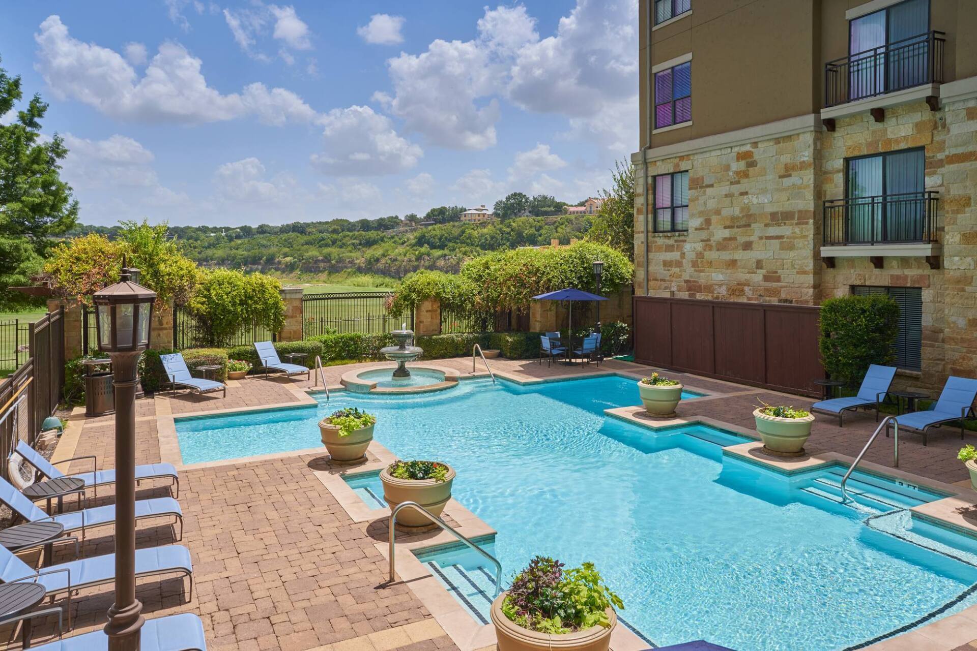 Pool area with loungers, fountain, and building in the background on a sunny day.