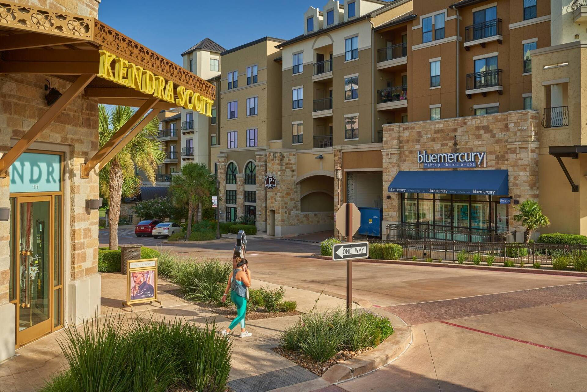 Streetscape with shops and apartments; person walking. Buildings are tan, brown, and beige.