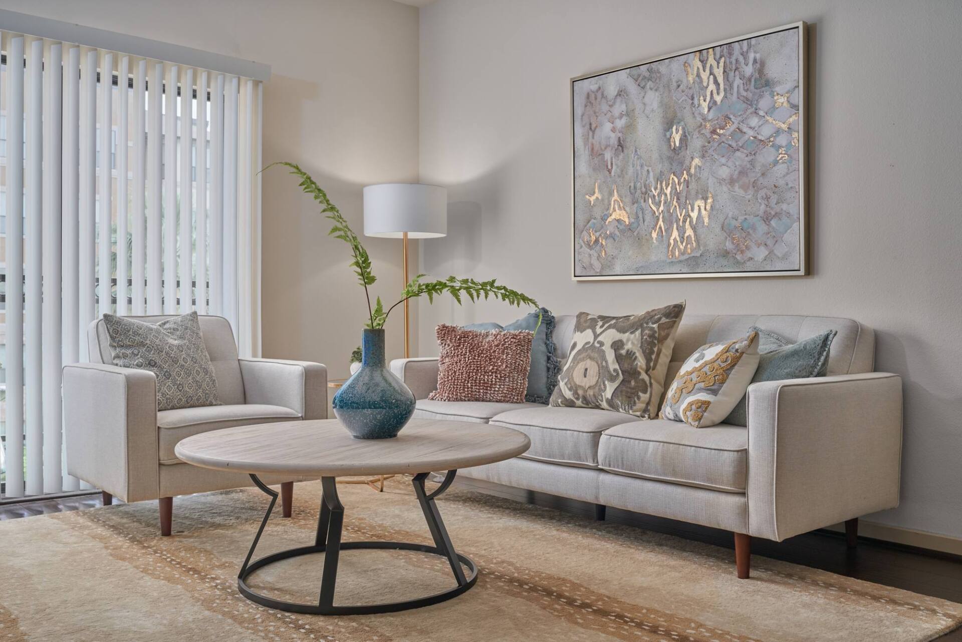 Living room with cream-colored furniture, rug, and artwork. A round coffee table and floor lamp are also present.
