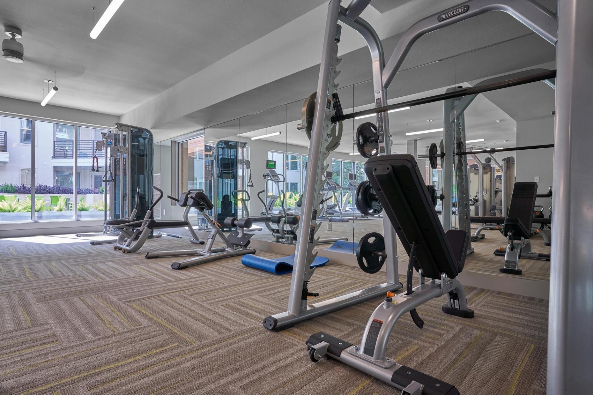 Gym with exercise machines: weight bench, stationary bikes, weight machines, mirrors, and windows.