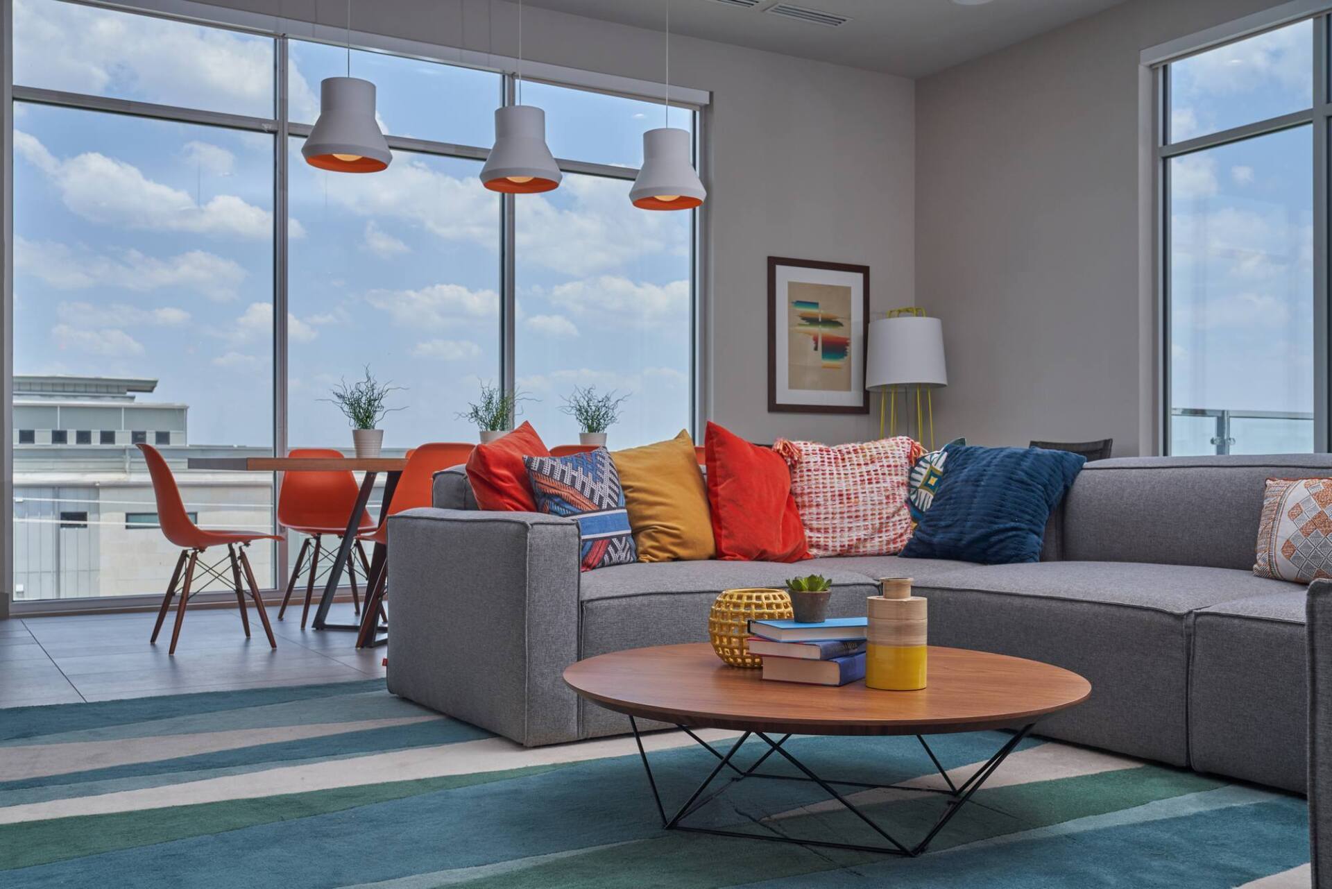 Living room with a gray sectional sofa, colorful pillows, round coffee table, and large windows with cityscape views.