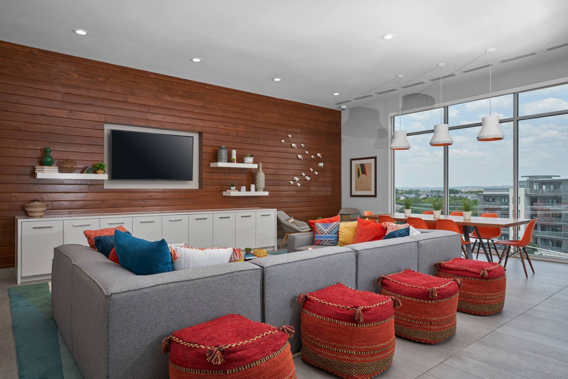 Modern lounge area with gray sectional sofa, red poufs, wood accent wall, and city view.