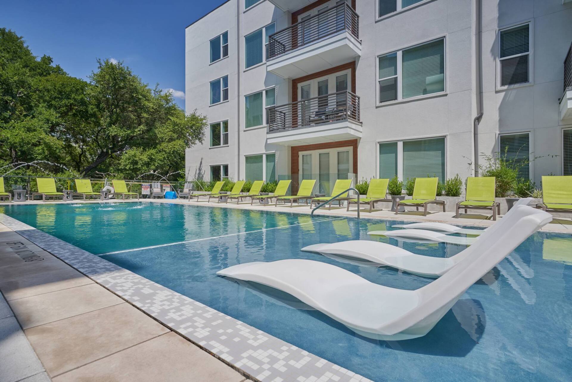 Swimming pool with white chaise lounges in water and green lounge chairs on the side of a modern building.