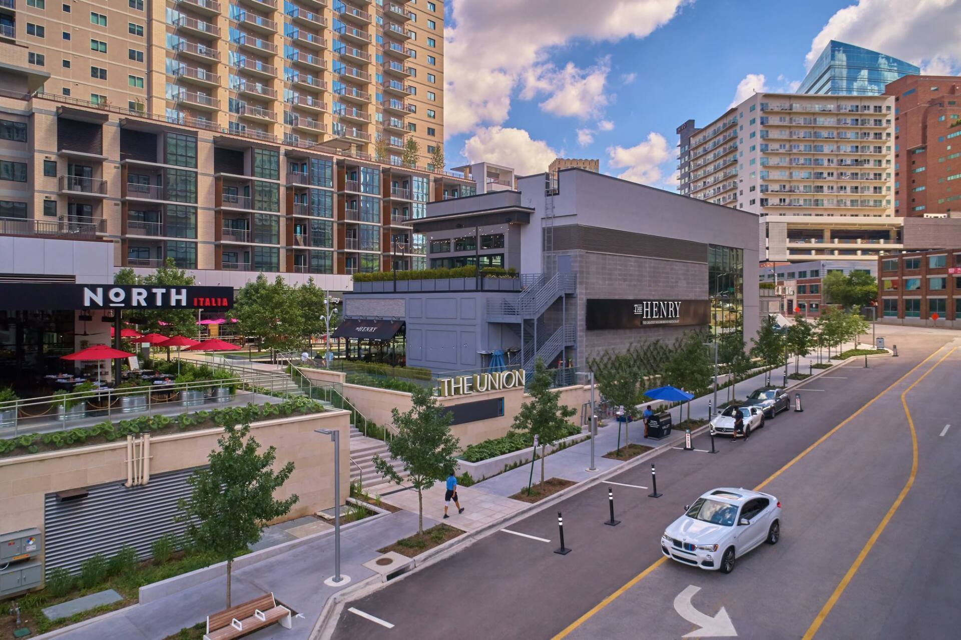 Street view with buildings, restaurants, a park with trees, and a car on the road.