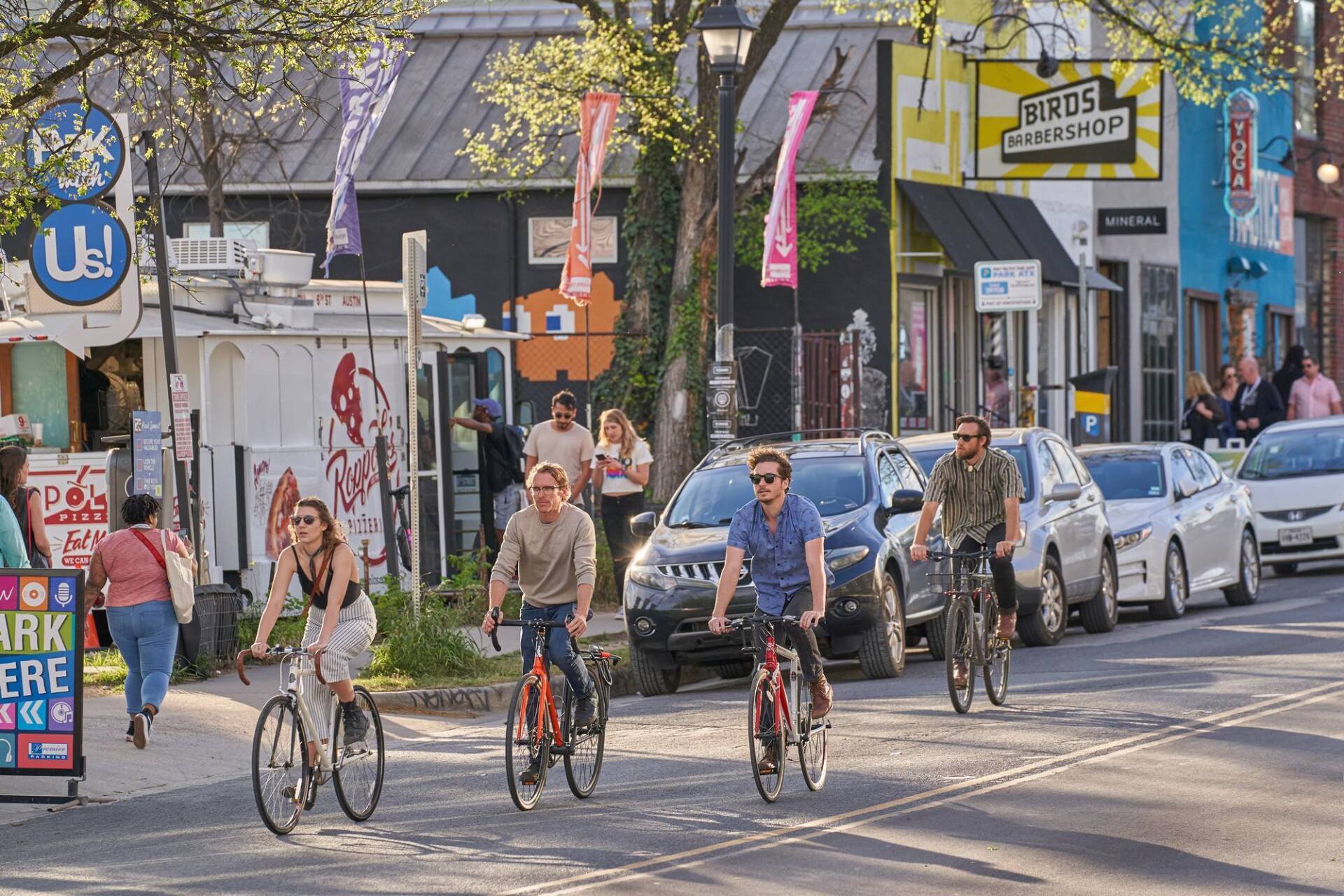 People biking down a city street with shops and parked cars on either side.