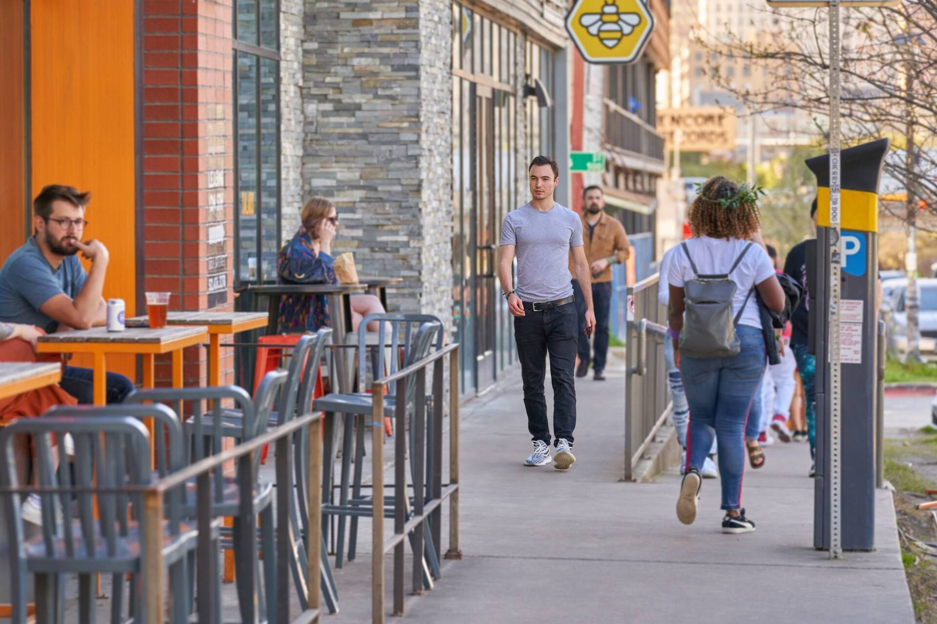 People on a city sidewalk, some walking, others at outdoor tables of a restaurant.