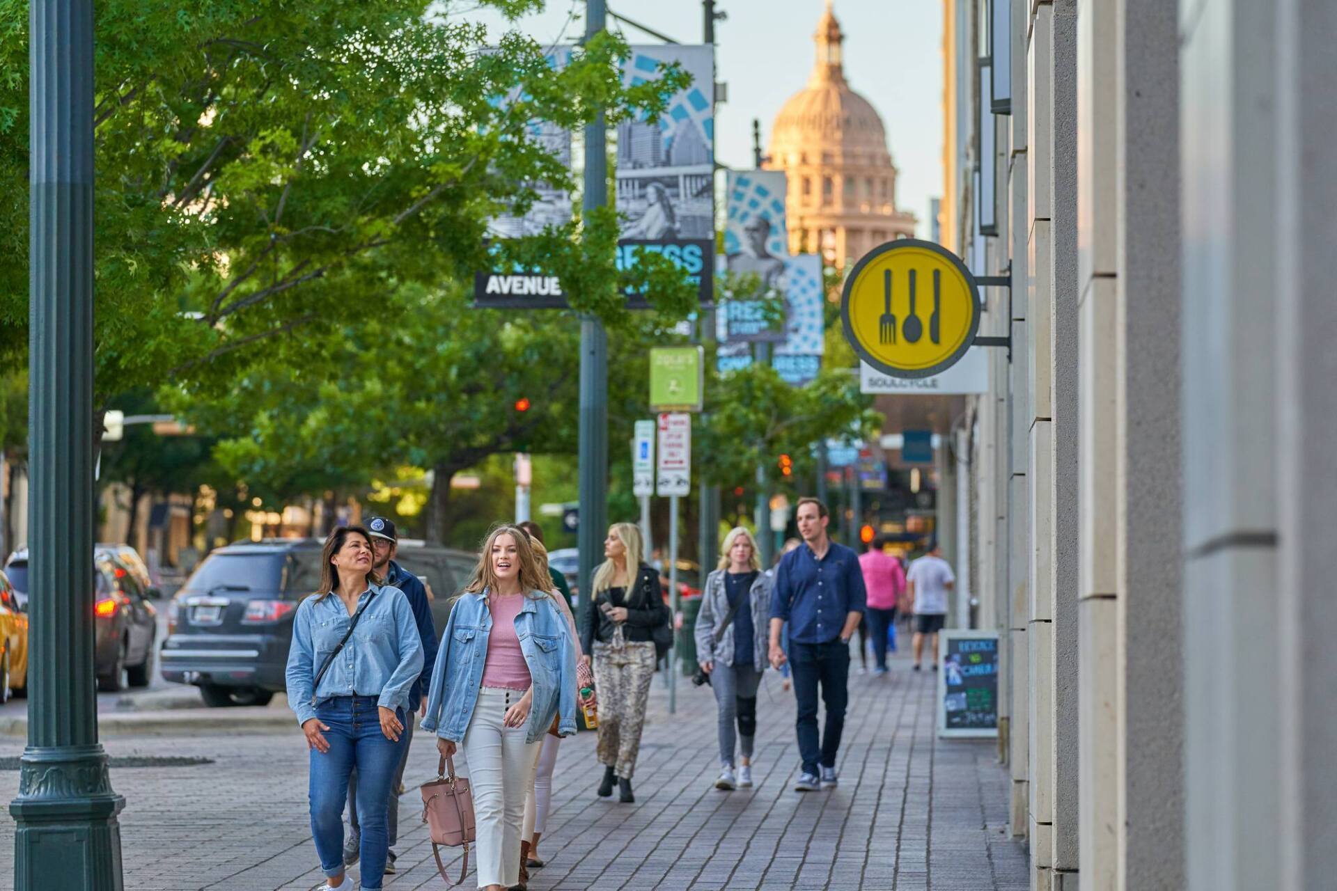 People walk on a sidewalk in Austin, TX, past shops, the Capitol, and street signs under a bright sky.