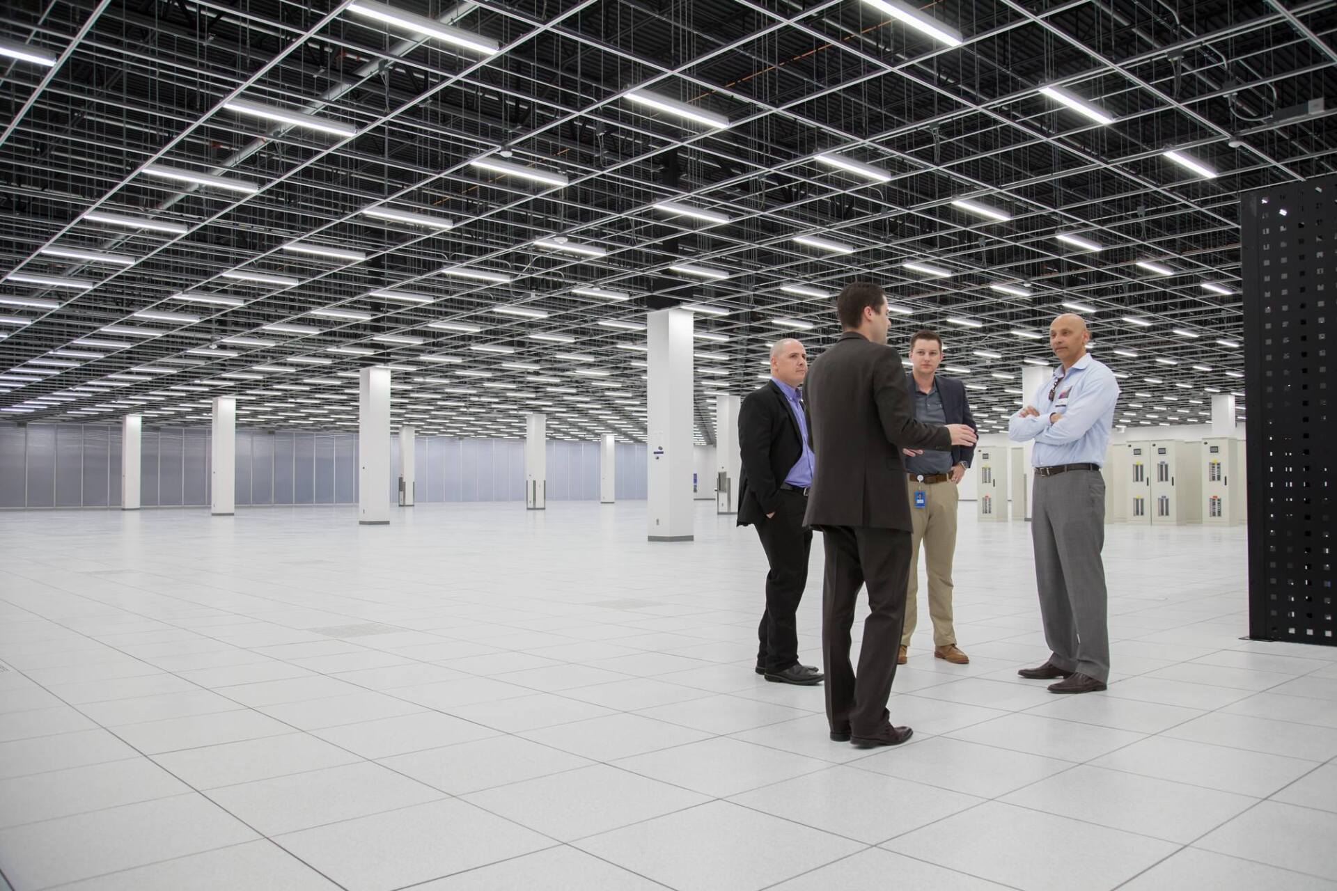 Four men in suits and business casual attire stand in a large, empty data center.