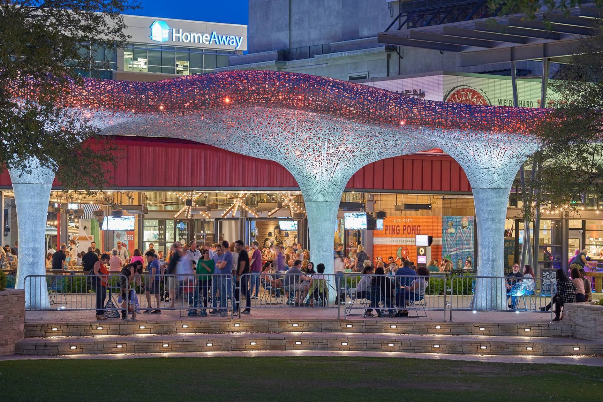 Outdoor patio with crowd under a decorative canopy lit with string lights. Restaurants and HomeTown Bank visible.