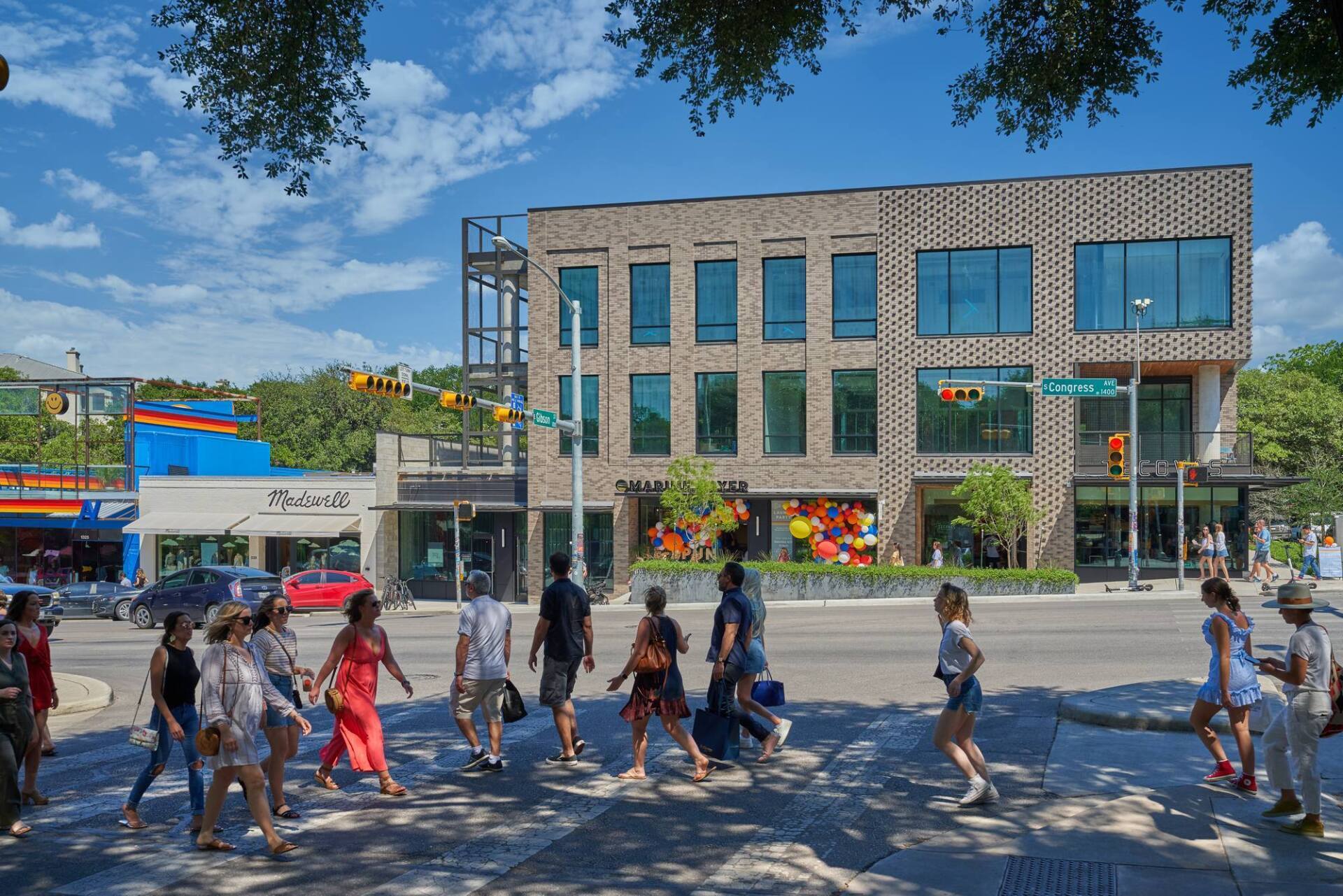 People crossing a street in front of a modern brick building and shops on a sunny day.