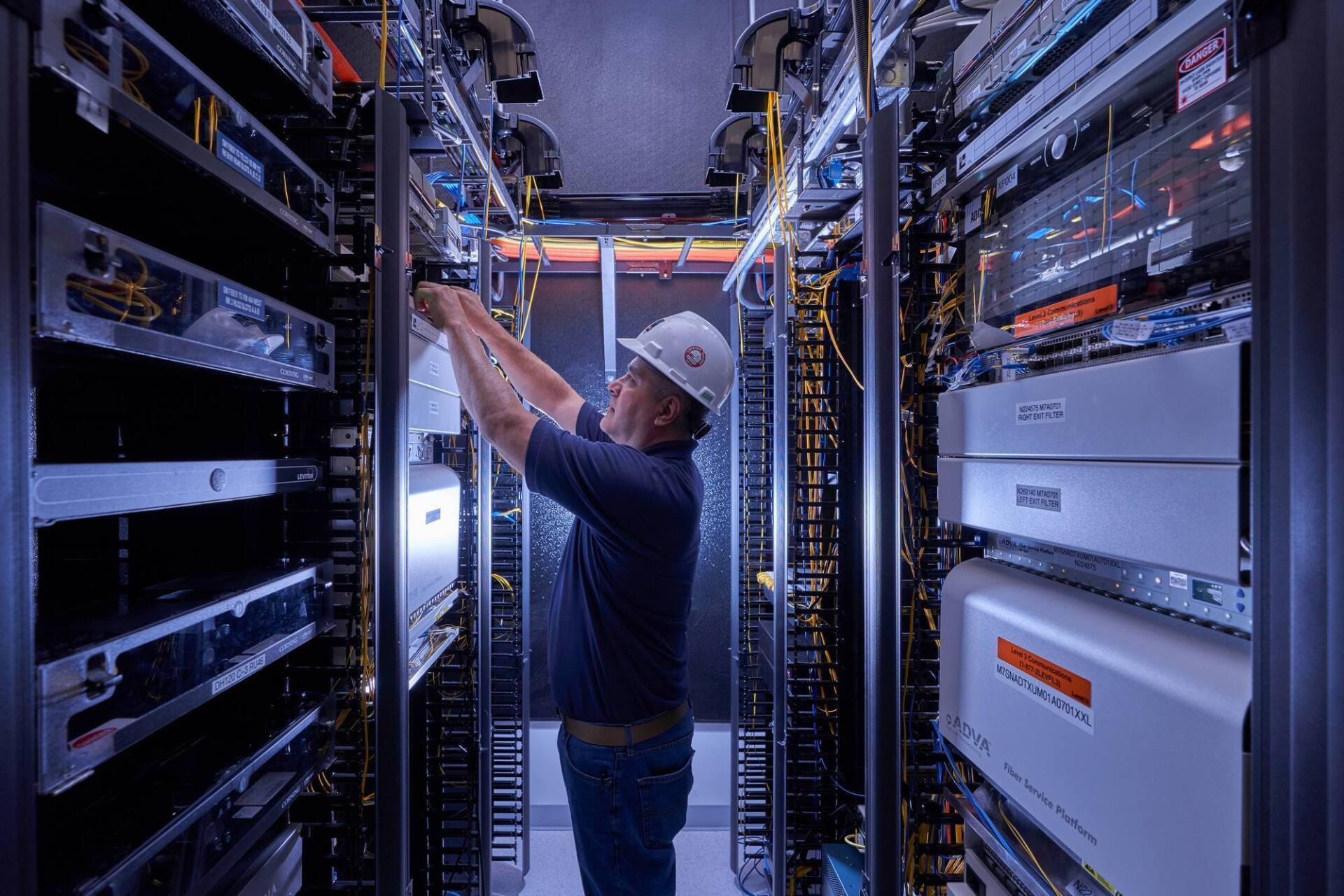 Man in a hard hat working on servers in a data center, surrounded by cables and equipment.