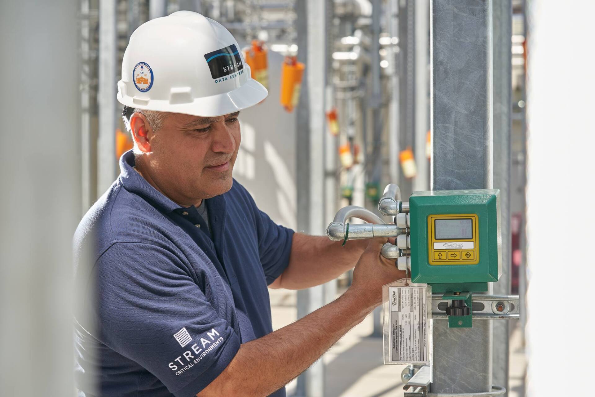 Man in hard hat examines industrial equipment outdoors. Green box on pipes.
