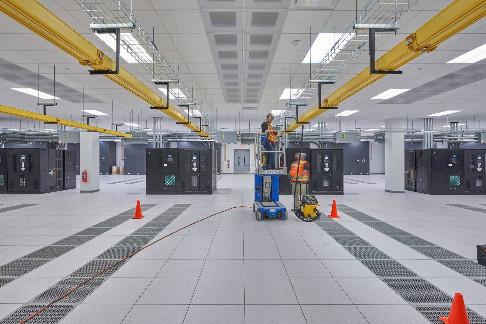 Workers in a large data center installing equipment using a lift. Yellow cable trays run overhead.