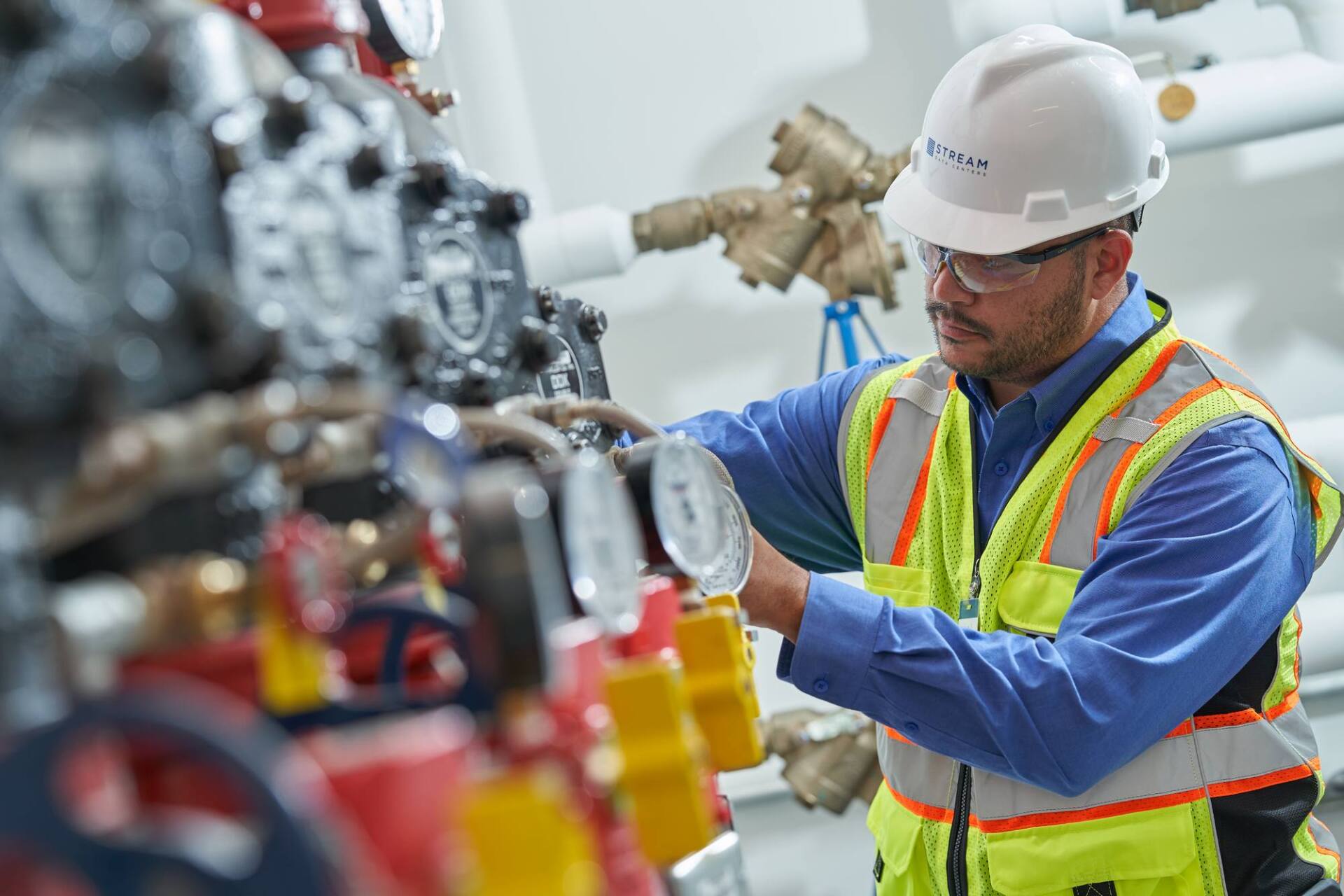 Man in hard hat and safety vest inspecting machinery in a mechanical room.