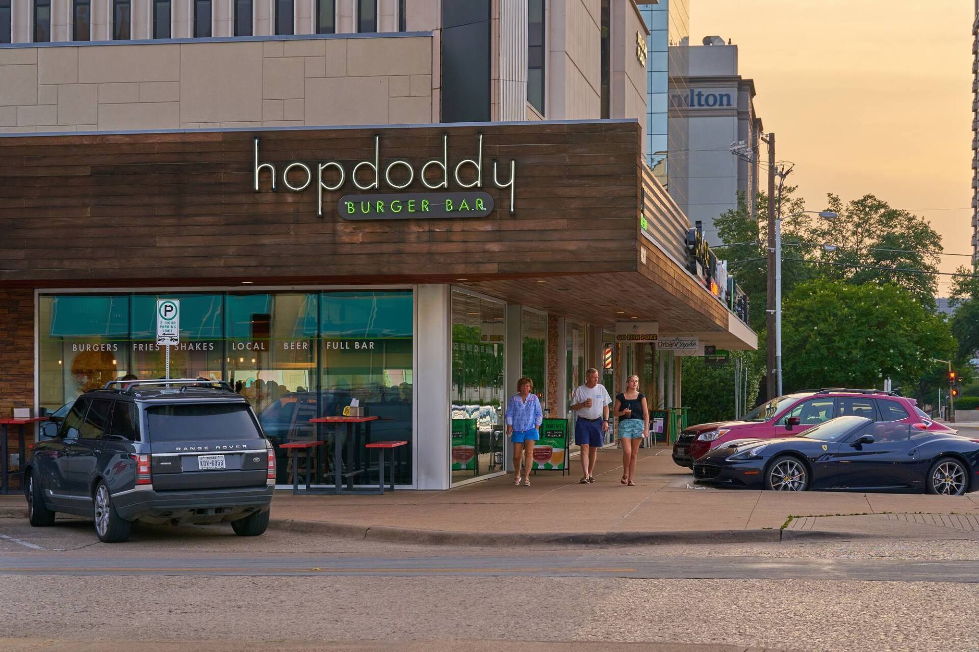 Hopdoddy Burger Bar storefront with people walking by, parked cars, and a building in background.