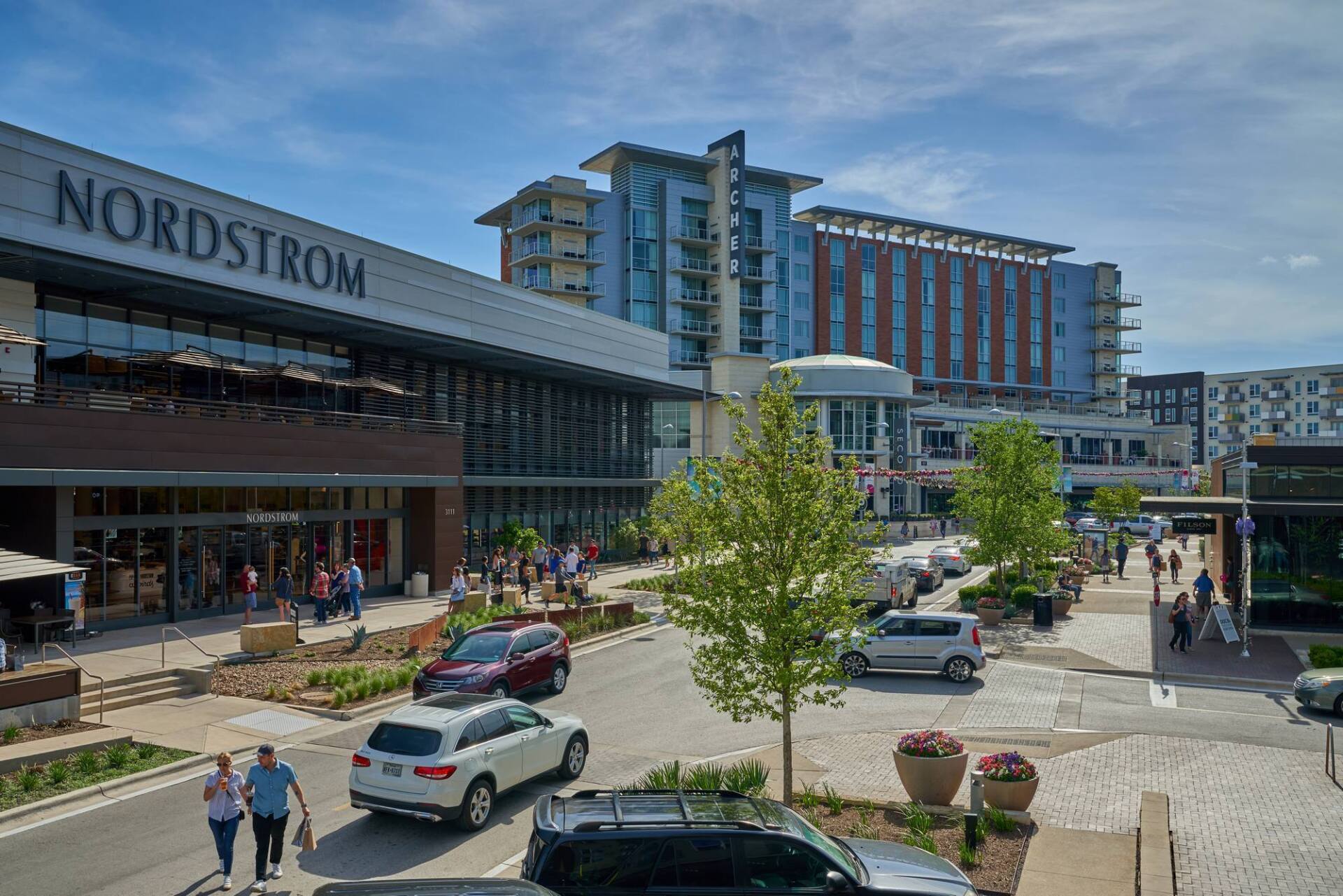 Nordstrom store and other buildings at outdoor shopping center, cars on street, pedestrians walking.