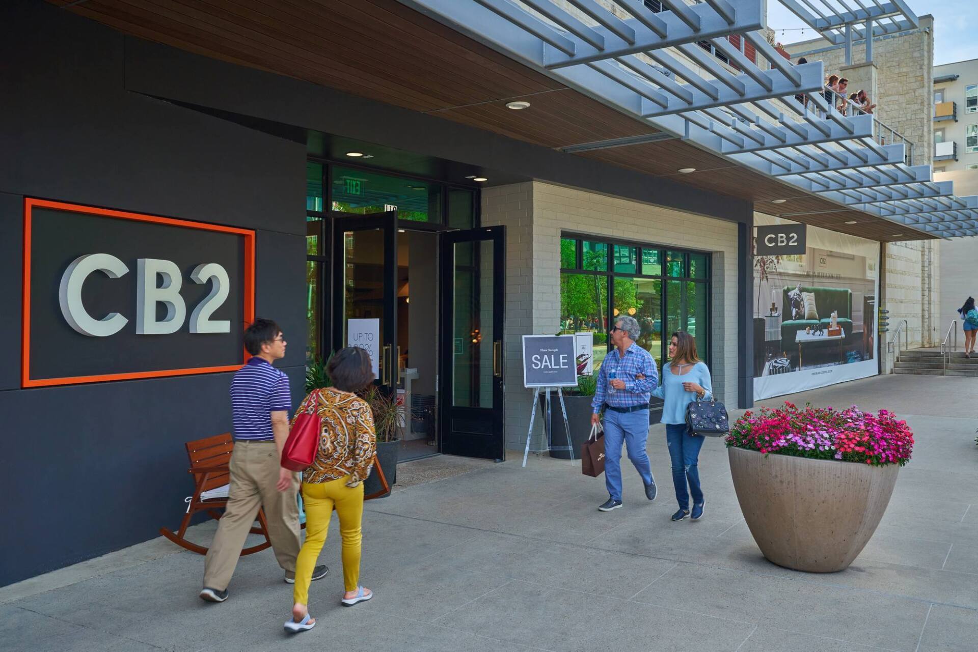 People entering CB2 store; exterior shot. Storefront has black exterior with a sign. Sidewalk and outdoor seating visible.