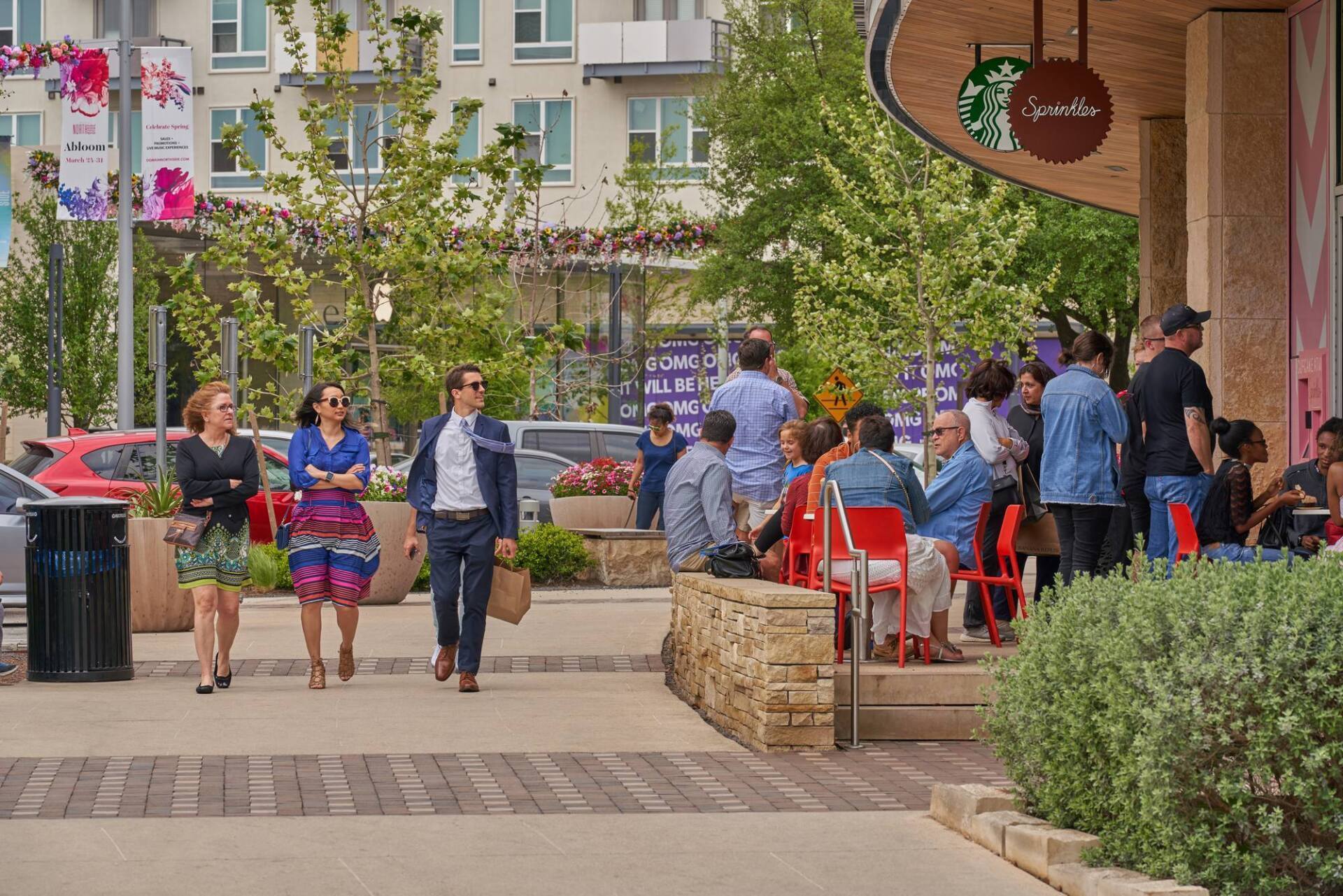People walking along a sidewalk, near a Starbucks with outdoor seating and trees.