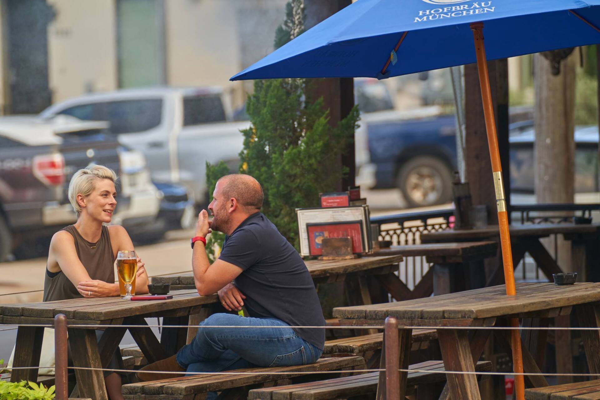 Two people seated at outdoor table with drinks, under an umbrella, smoking, and talking.