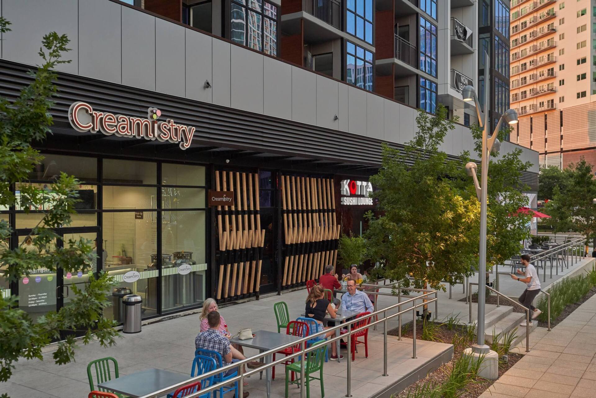 Restaurant storefront with outdoor seating; people dining. Building facade, vertical wood slats, trees.