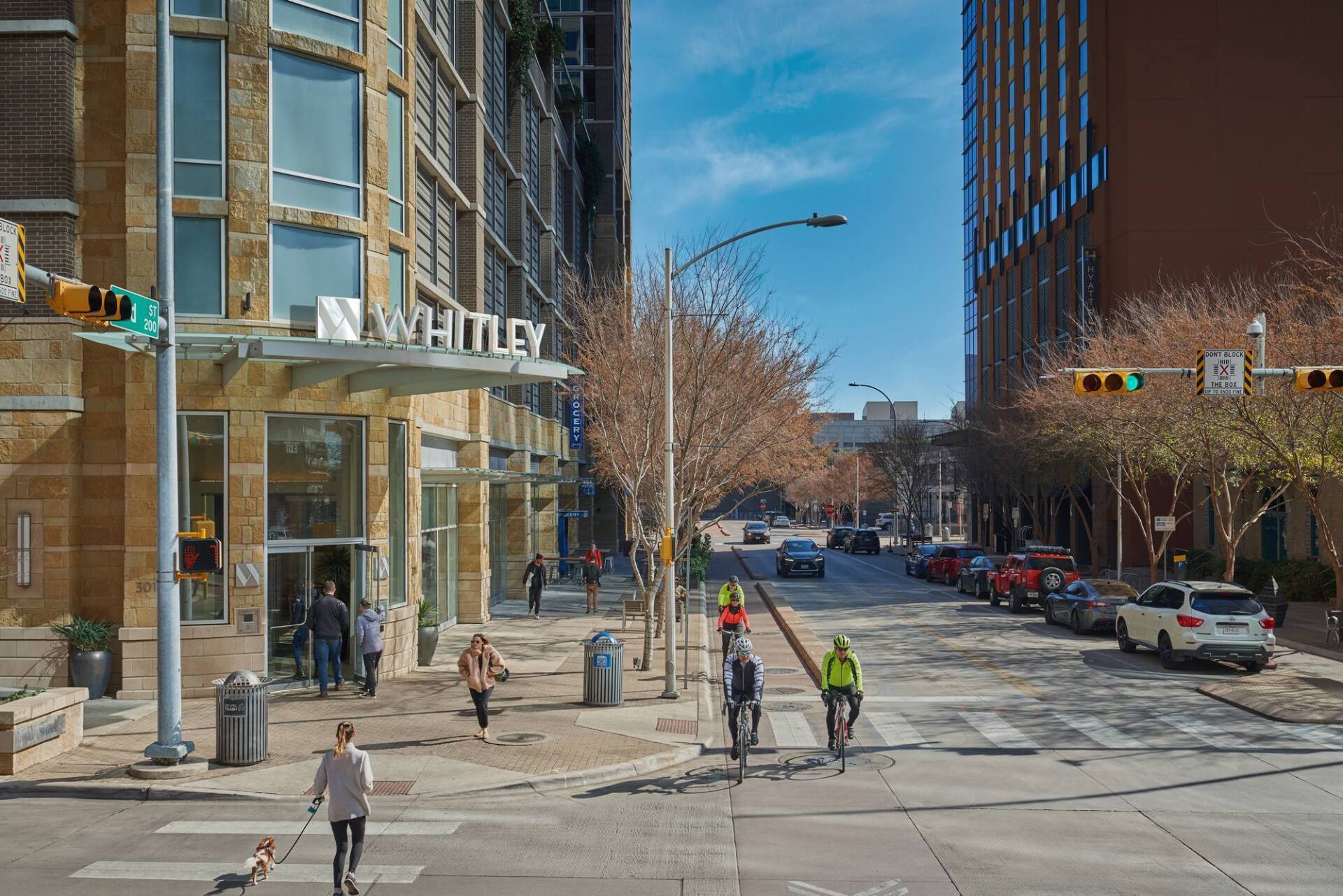 Street scene with buildings, pedestrians, cyclists, and traffic lights. Sign reads 