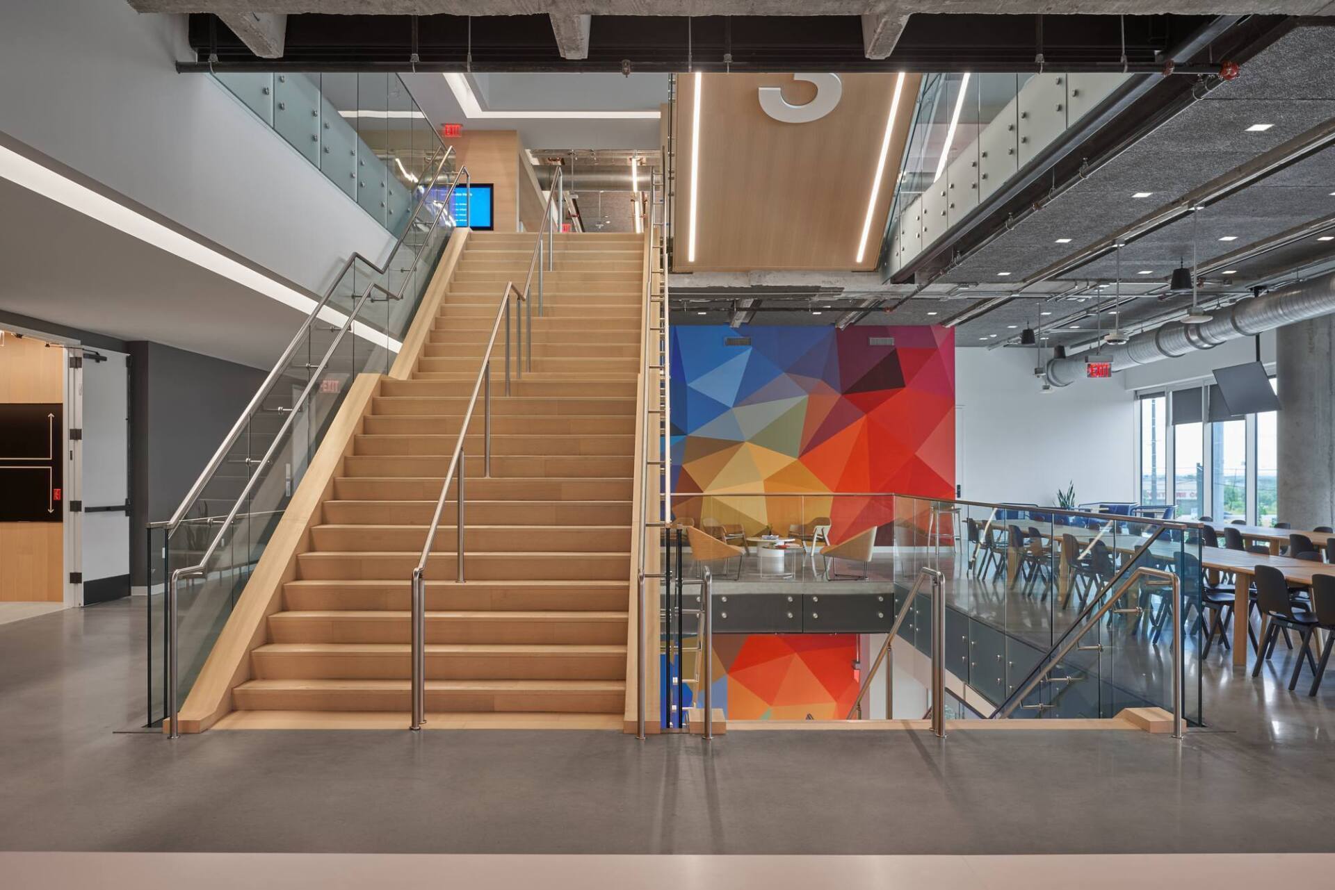 Staircase with wooden steps and glass railings, leading to upper level with seating area and colorful geometric wall art.