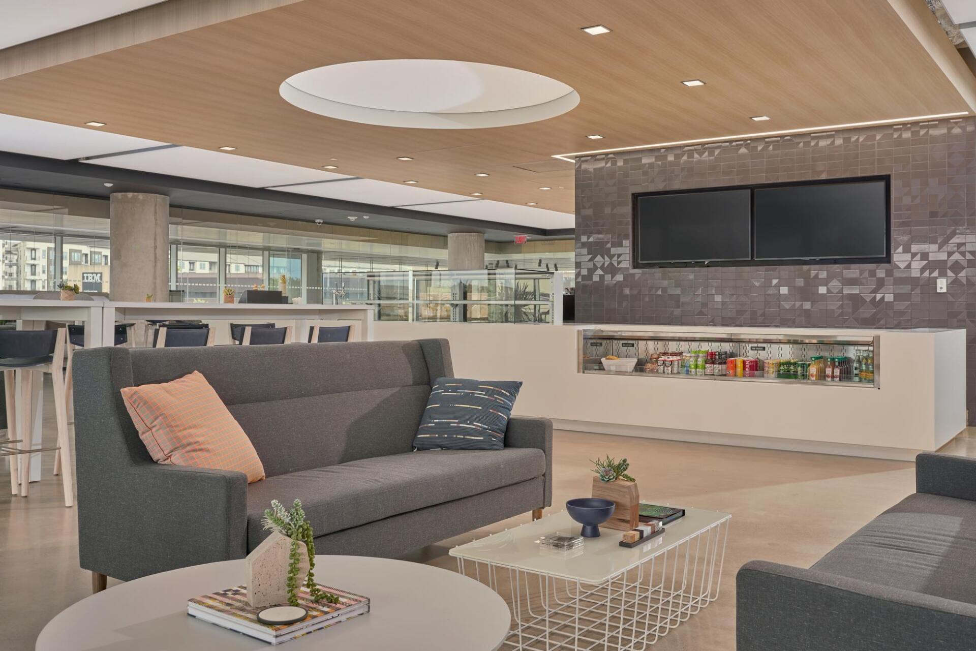 Modern lounge area with gray sofas, low tables, and a refreshment counter with TVs.