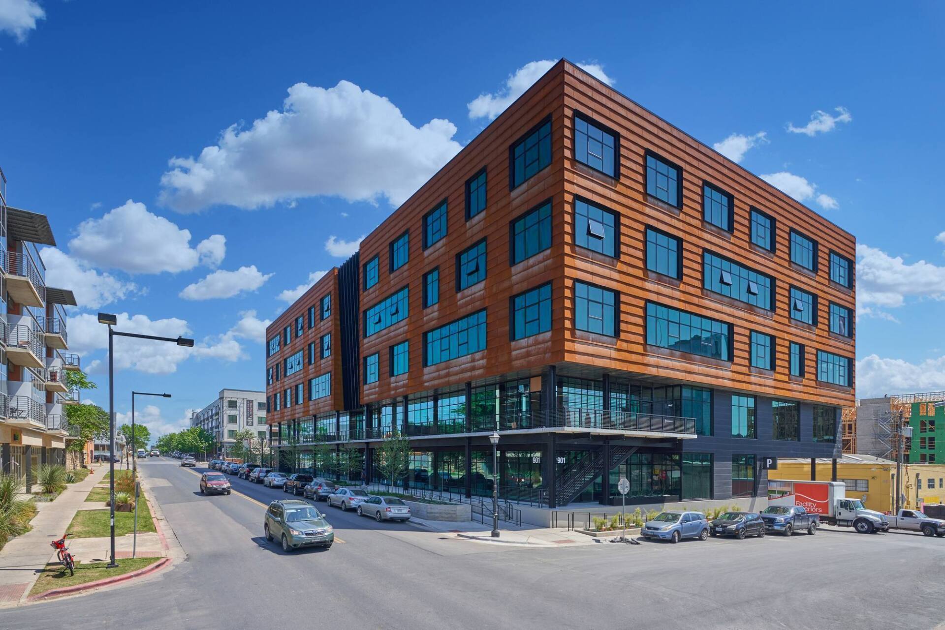 Modern copper-paneled building with large windows on a sunny street, parked cars, and a blue sky.