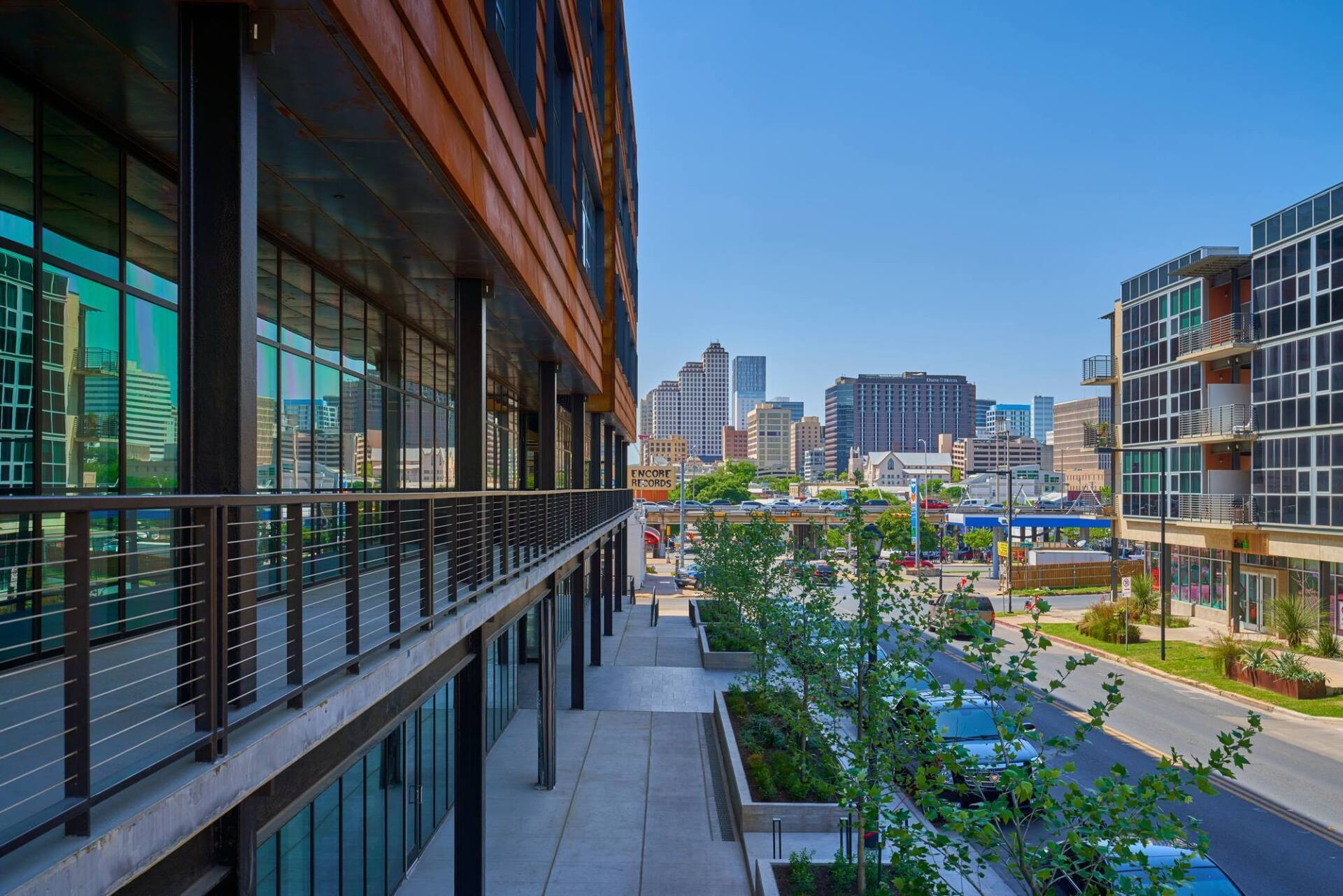 Modern buildings with glass windows and a balcony overlook a city street with distant skyscrapers.