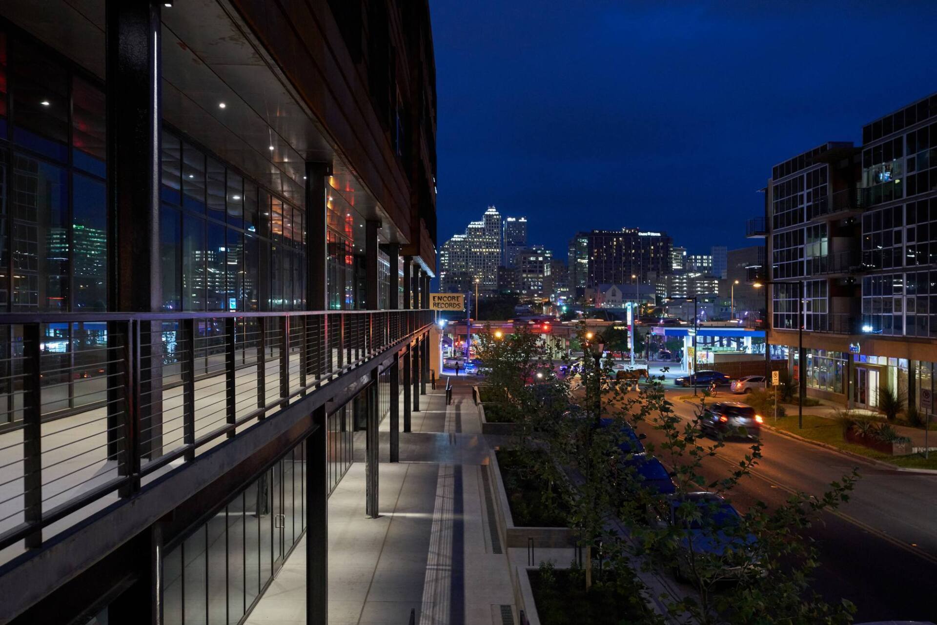 Night view of a city street with illuminated buildings, a walkway, and cars driving, under a dark blue sky.