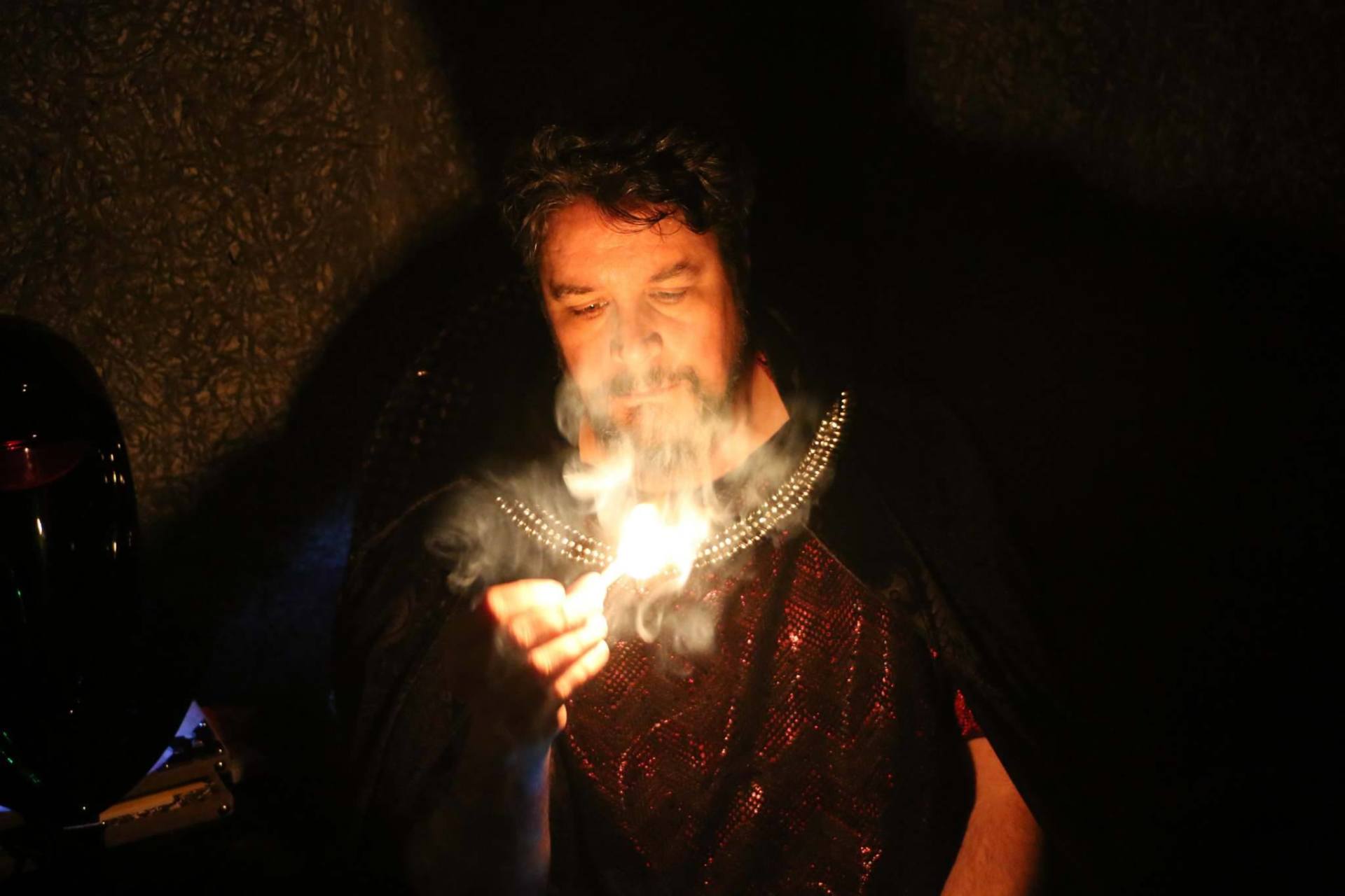 A man wearing a cape and holding a flame, surrounded by the dark, representing Hades.