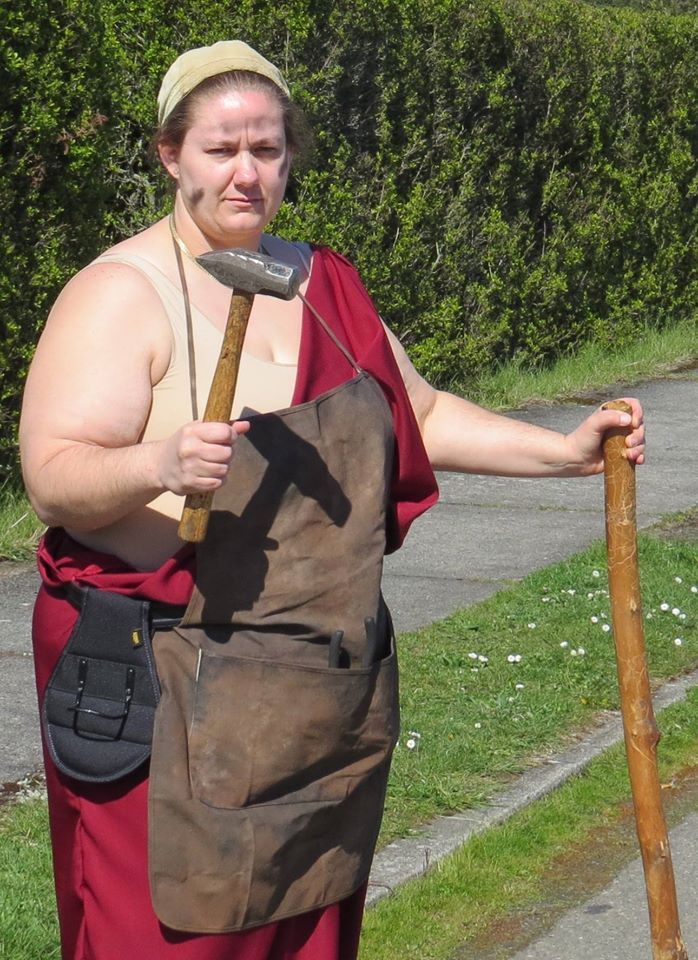 A woman in a red robe is holding a hammer and a stick,   representing Hephaestus.
