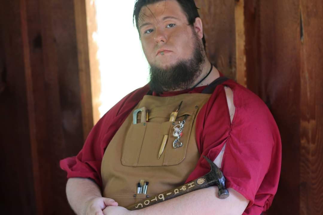 A man with a beard is wearing an apron and holding a hammer, representing Hephaestus.