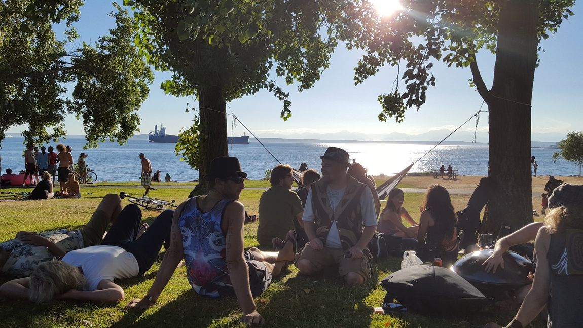 A group of people are sitting in the grass near the water, gathering for the Aquarian Tabernacle Church.