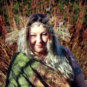 A woman wearing a headband and a scarf is standing in front of a bush, representing Demeter.