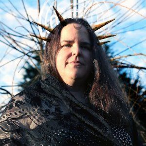A woman wearing a crown of horns is standing in front of a tree, representing Hekate.