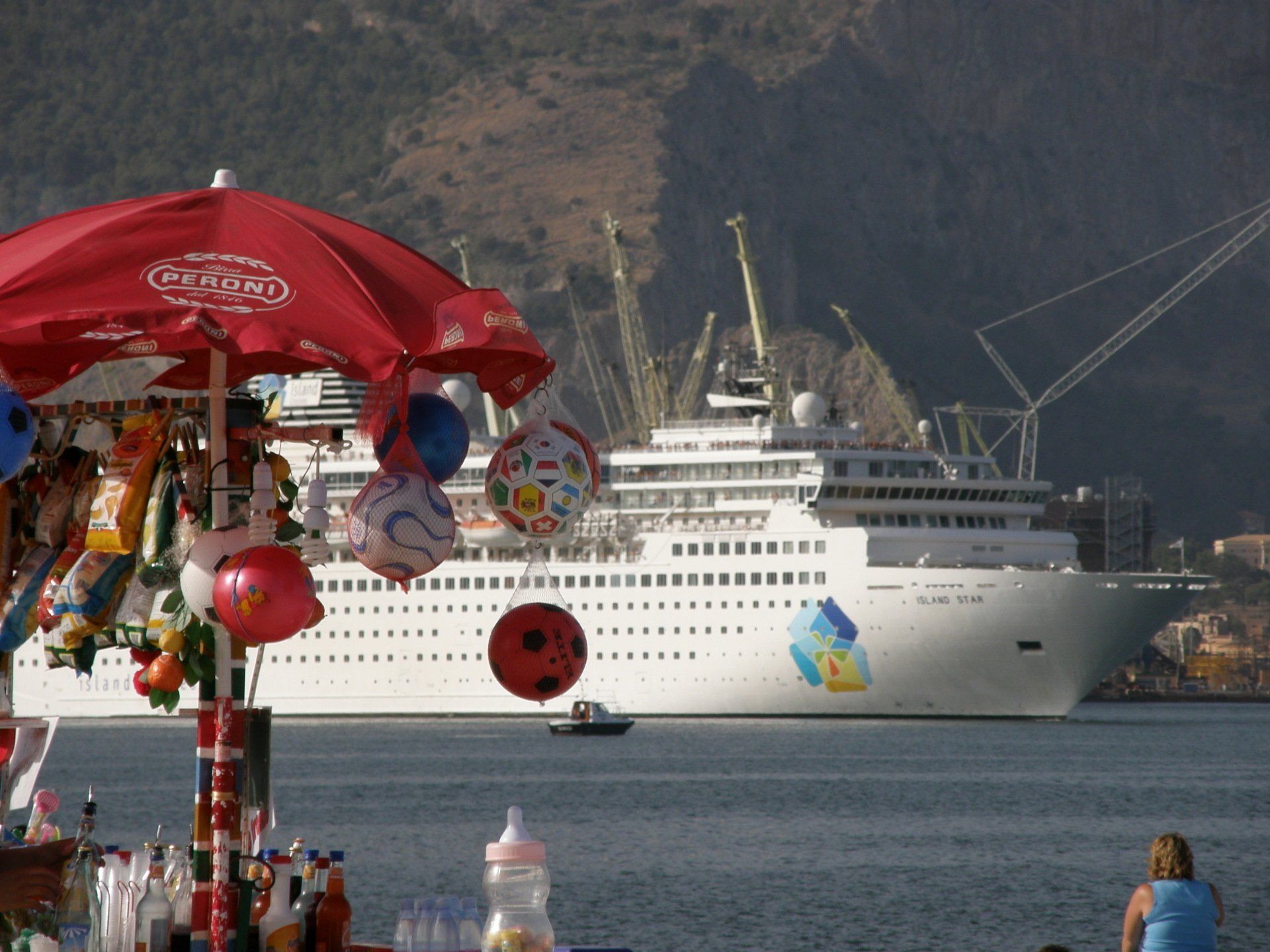 Ein Kreuzfahrtschiff im Hafen hinter einem roten Souvenirstand, im Hintergrund Berge.