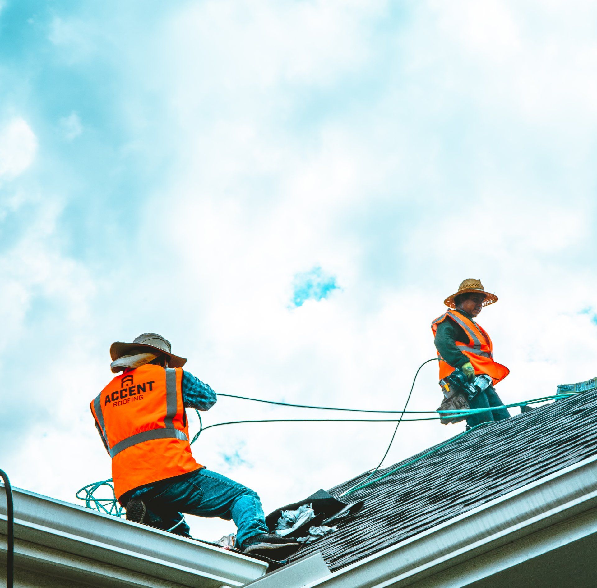 Two roofers in orange vests and hats on a roof working on a bright cloudy day.