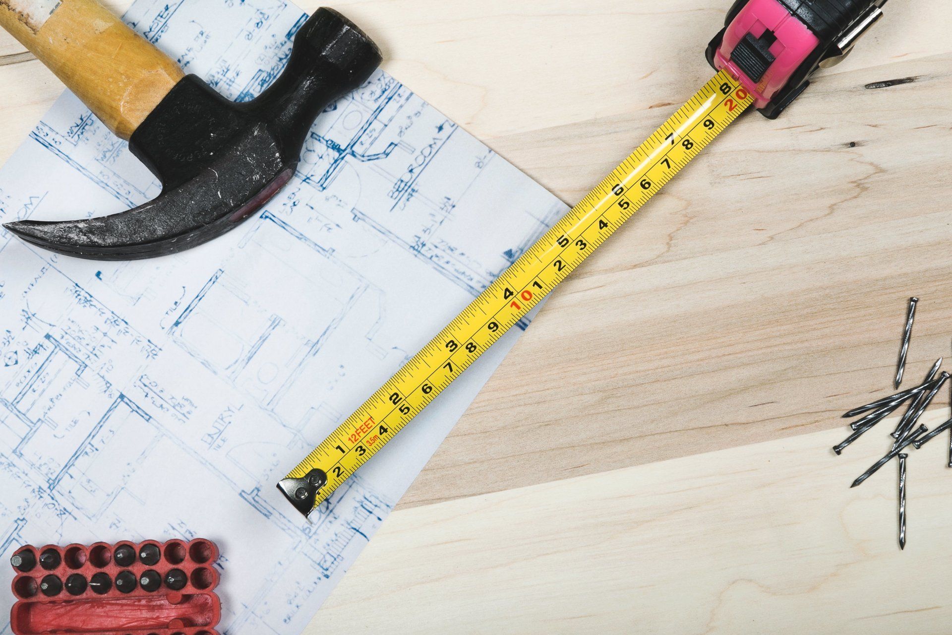 Hammer, measuring tape, and blueprints on a light wood surface.