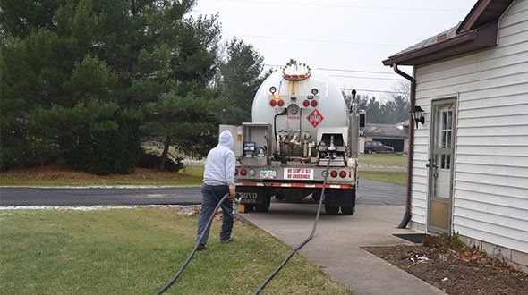 A propane truck refills a tank at a house, a person in a hoodie pulls a hose.
