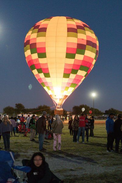 Colorful hot air balloon illuminated against a twilight sky, people gathered on a grassy field.