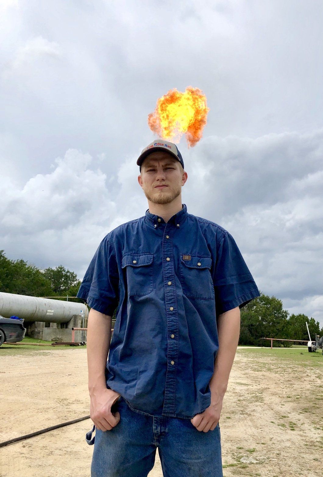 Man in denim shirt and hat with fire over his head, standing outside.