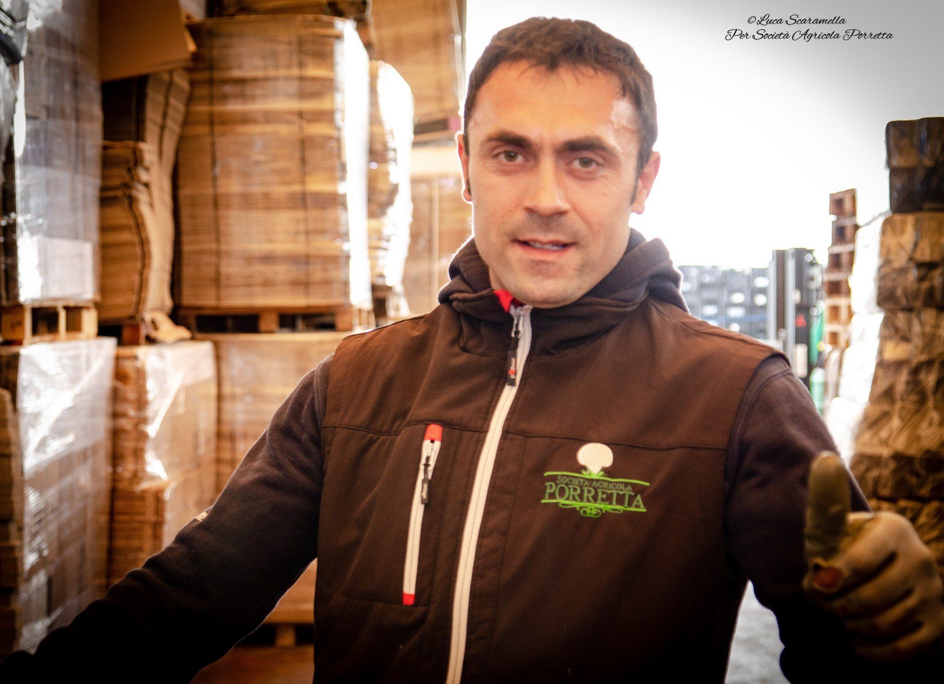 Un uomo con il gilet alza il pollice, in piedi in un magazzino con materiali accatastati.