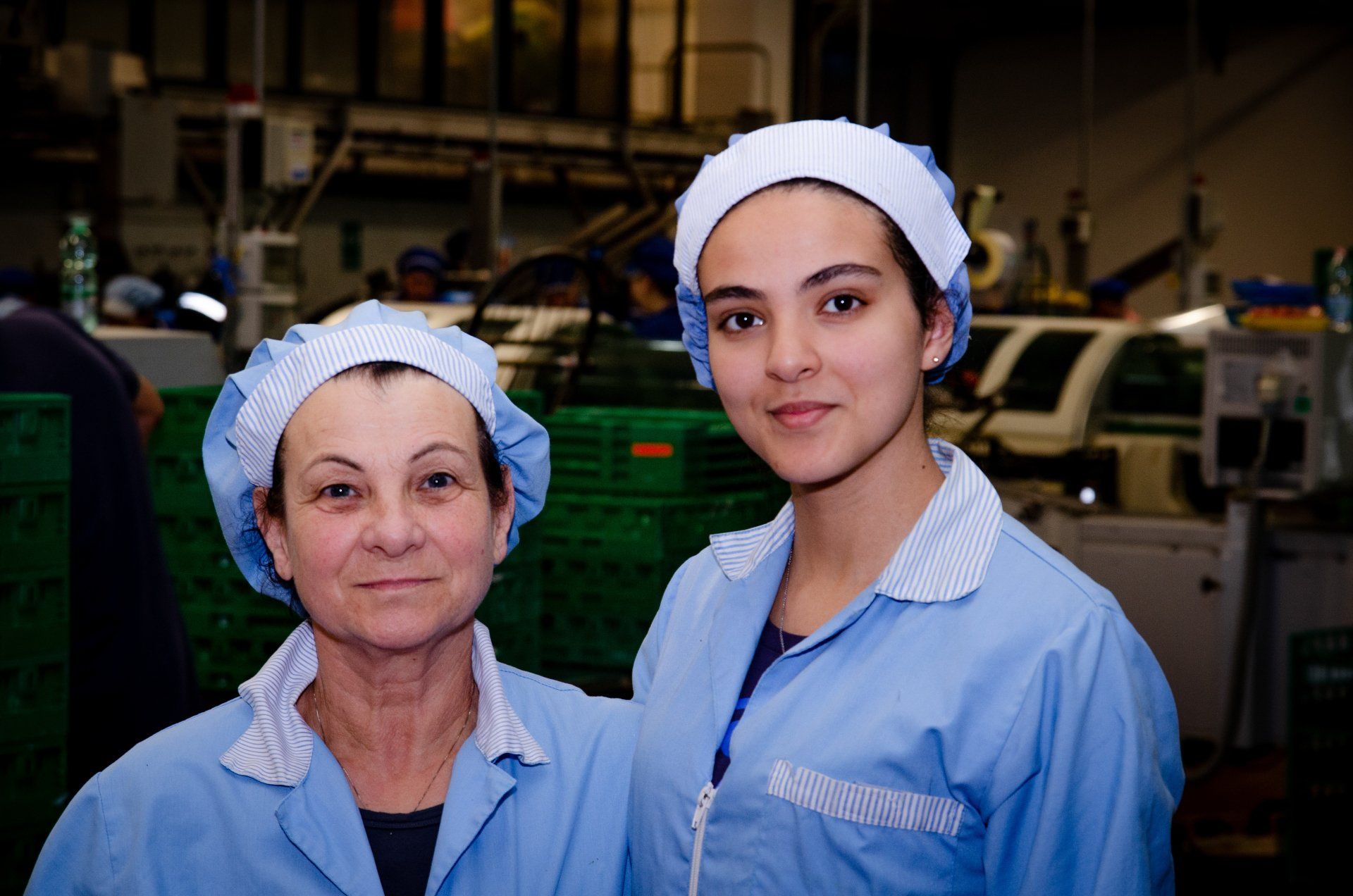 Due persone che indossano uniformi da lavoro azzurre e retine per capelli sorridono in una fabbrica.