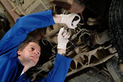 Mechanic in blue coveralls inspecting the underside of a car with a wrench