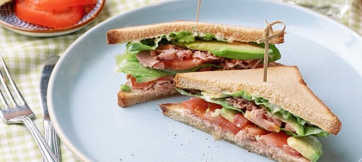 Twee driehoekig gesneden avocado- en rosbiefsandwiches op een lichtblauw bord, met een vork en plakjes tomaat ernaast.