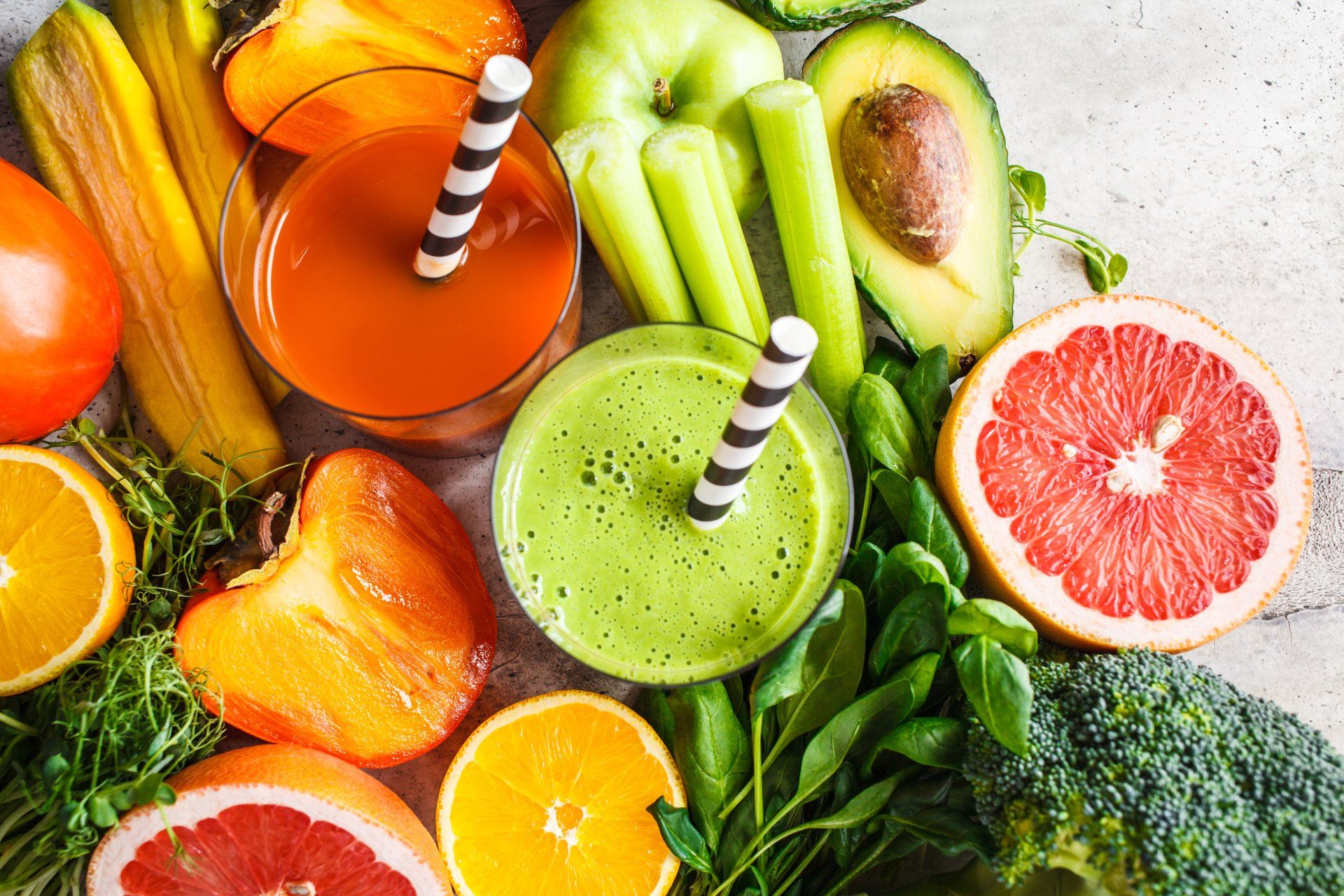 Twee glazen sap – een sinaasappelsap en een groen sap – omringd door verse groenten zoals grapefruit, avocado, selderij en broccoli.